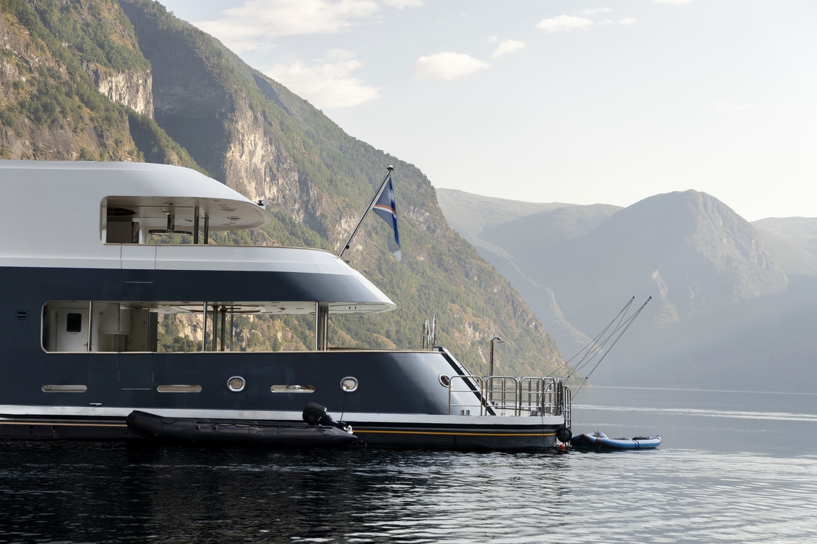 A side view of luxury expedition vessel Aqua Lares cruising past a green cliff.