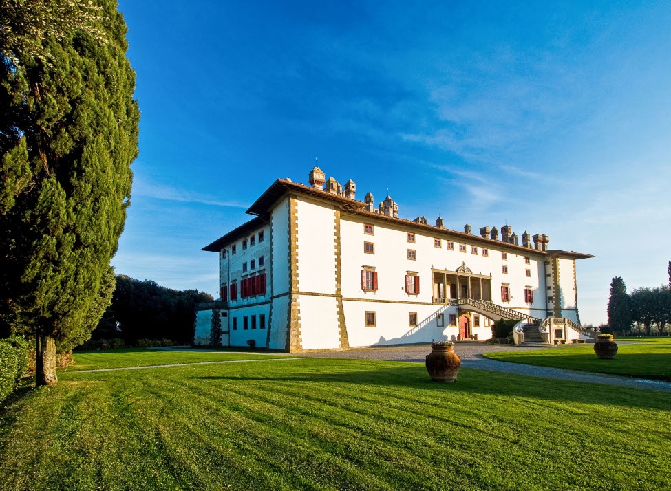 A large, white, castle-like villa stands on mowed lawn with several large decorative pots and a blue sky in the background.