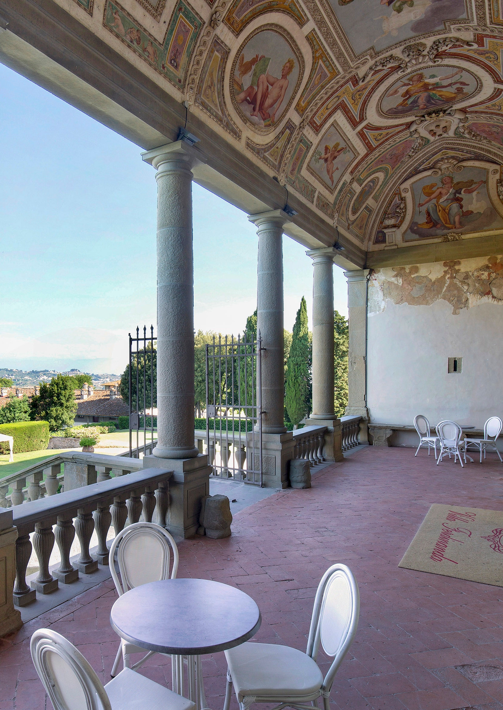 Terrace with a single table and two chairs sitting within a columned area with murals painted on the ceiling and a view out onto the countryside.