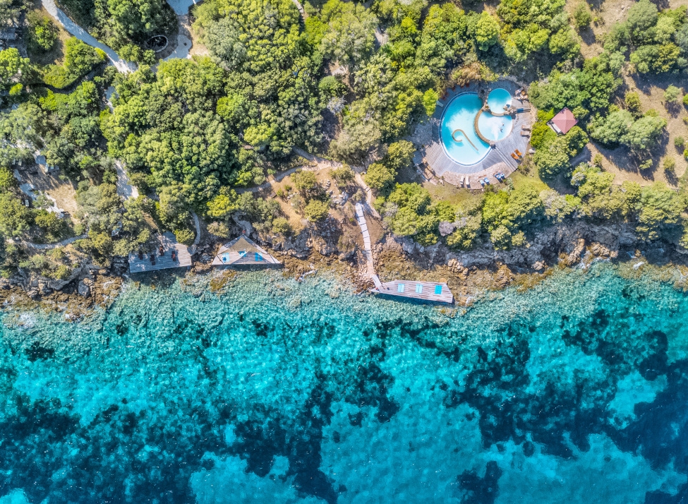 Aerial view of coast with clear turquoise water, green trees and a pool nestled into them.