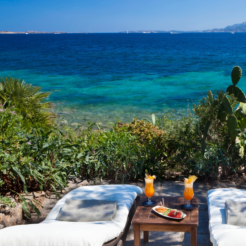 Two white lounge chairs with a table containing fruity drinks in between, overlooking greenery and a turquoise sea view.