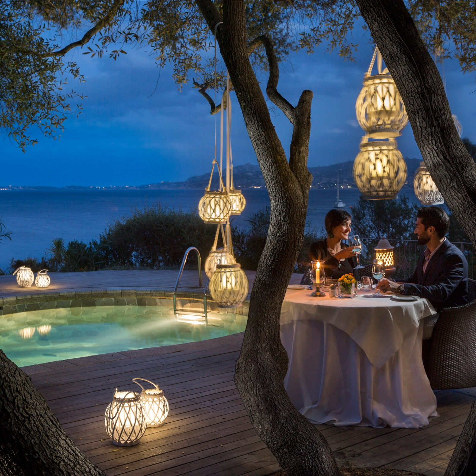 Evening scene of a people seated at an outdoor dining table surrounded by warmly lit lanterns, the pool and sea beyond.