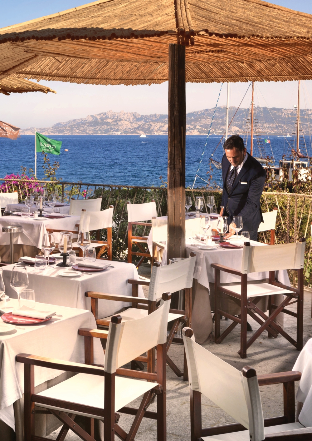 An outdoor dining area where a male waiter is setting an empty table under an umbrella, with the sea in the background.