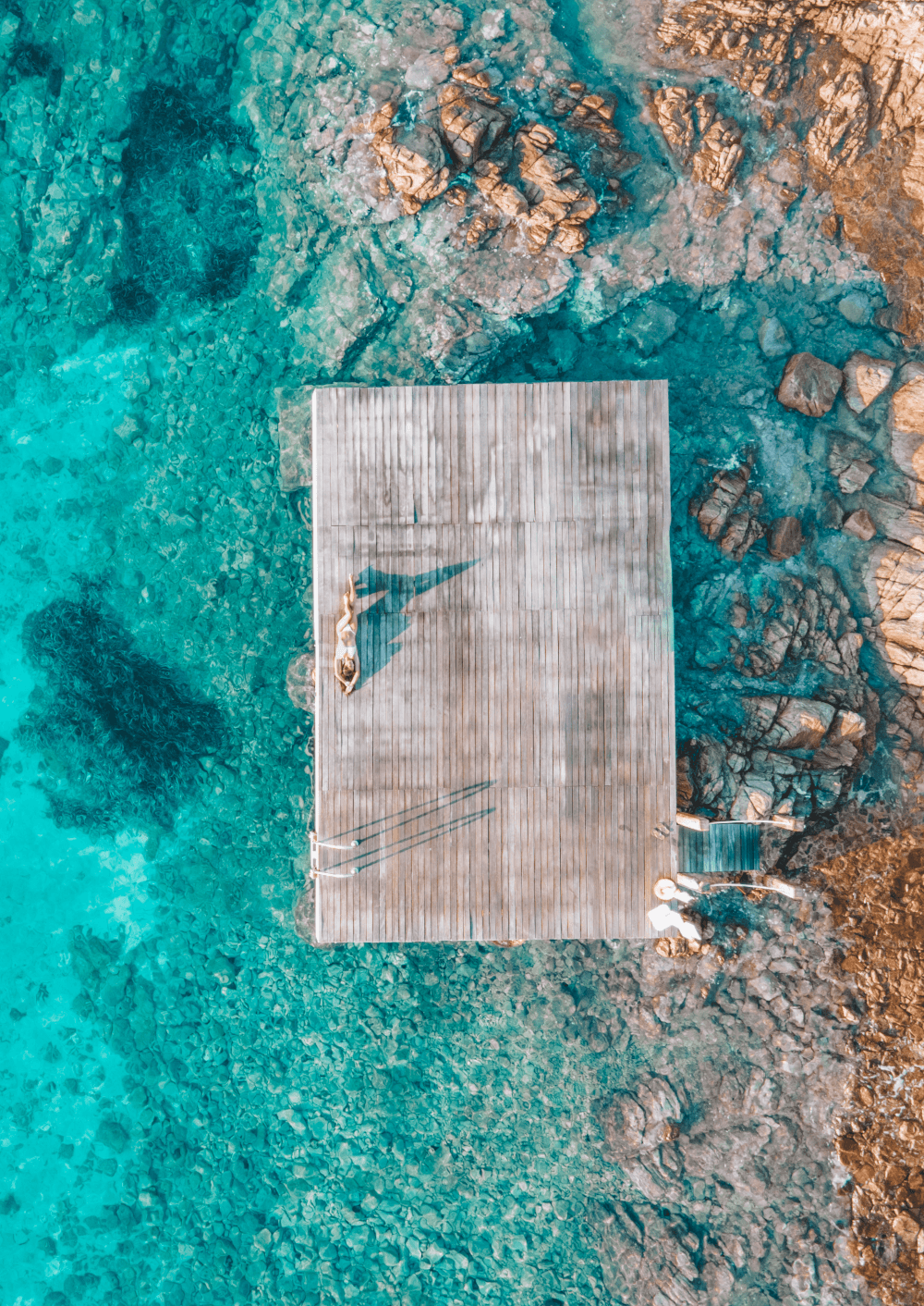Aerial view of a wooden floating dock on a turquoise sea with rust and brown rocks.