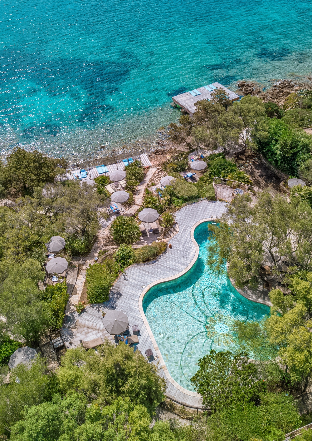 Aerial view of the pool, walkways and umbrellas set into greenery beside the tuquoise sea.