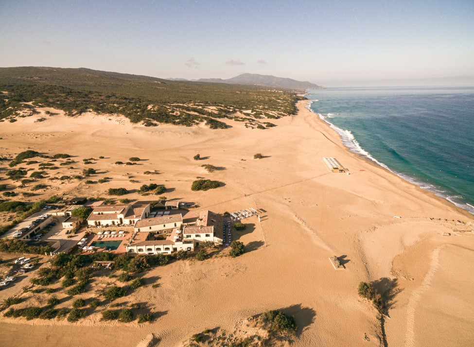 Aerial view of a big sandy beach with the sea on the right side and the property on the left tucked into some greenery.