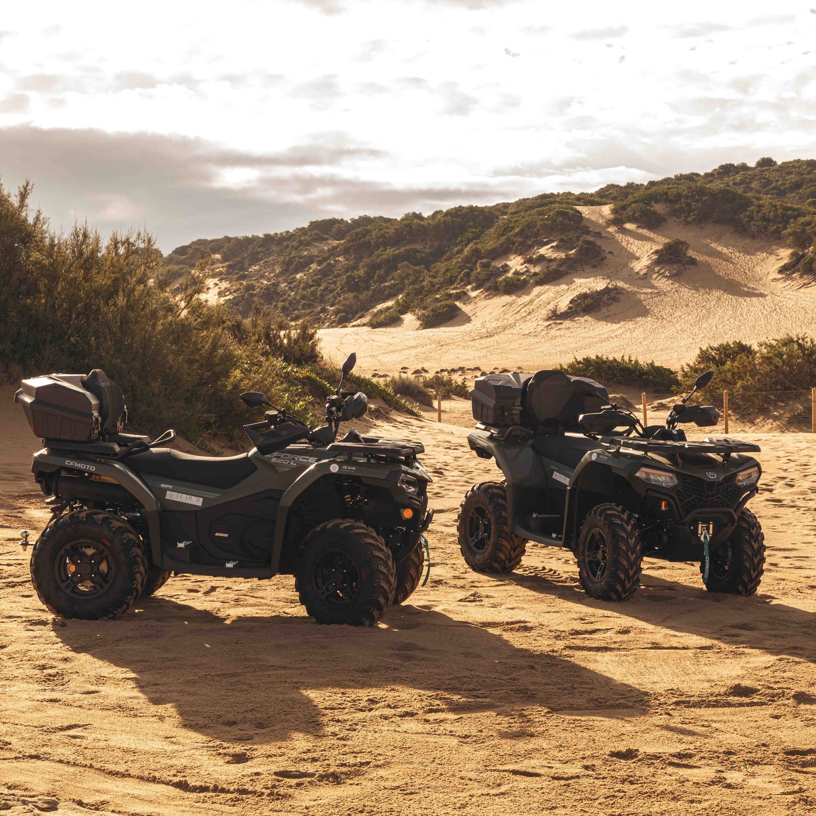 Two quadbikes sitting on a stretch of sand with dunes and greenery in the background.