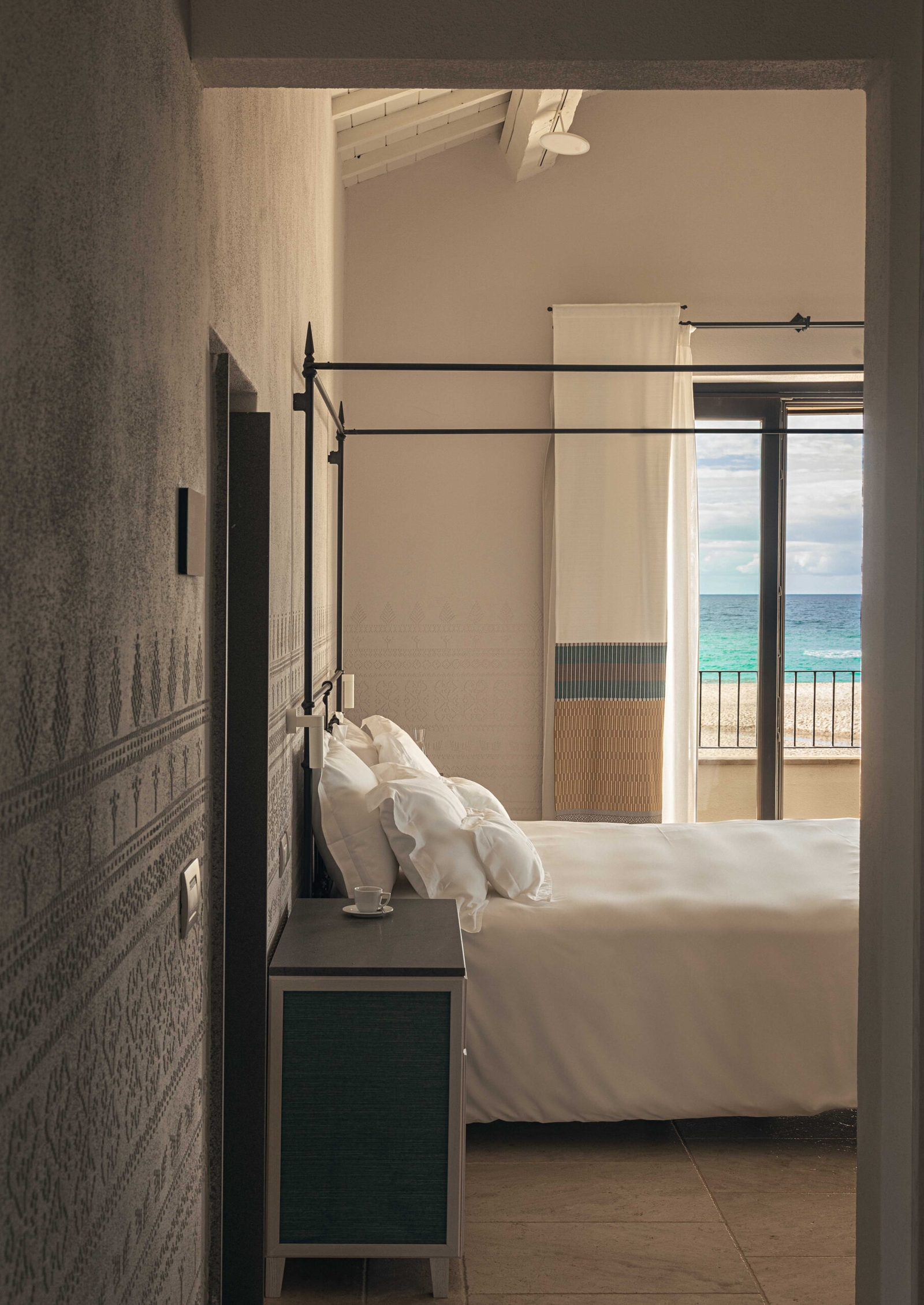 Interior of a bedroom looking through a doorway onto a bed with an open terrace door and turquoise sea in the background.