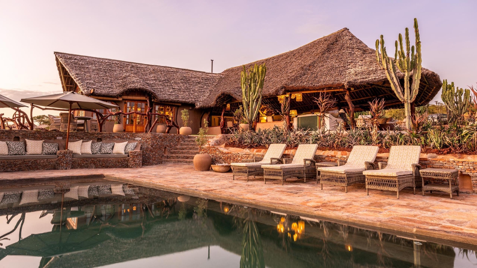 A luxury safari lodge with a swimming pool, lounge chairs, and thatched-roof buildings surrounded by desert plants.