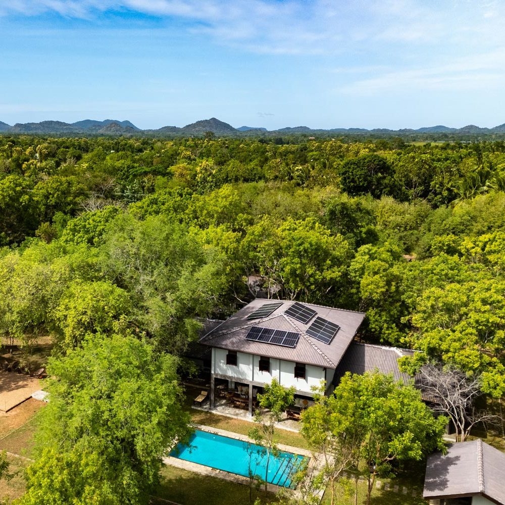 Drone view of a house with solar panels and a blue pool surrounded by a vast green forest.