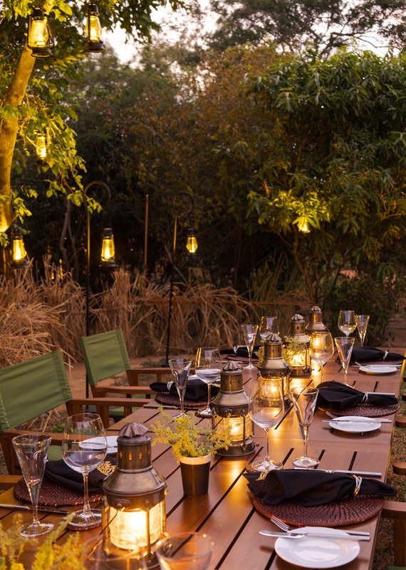 A wooden outdoor table set with wine glasses and lanterns under trees decorated with hanging lights.