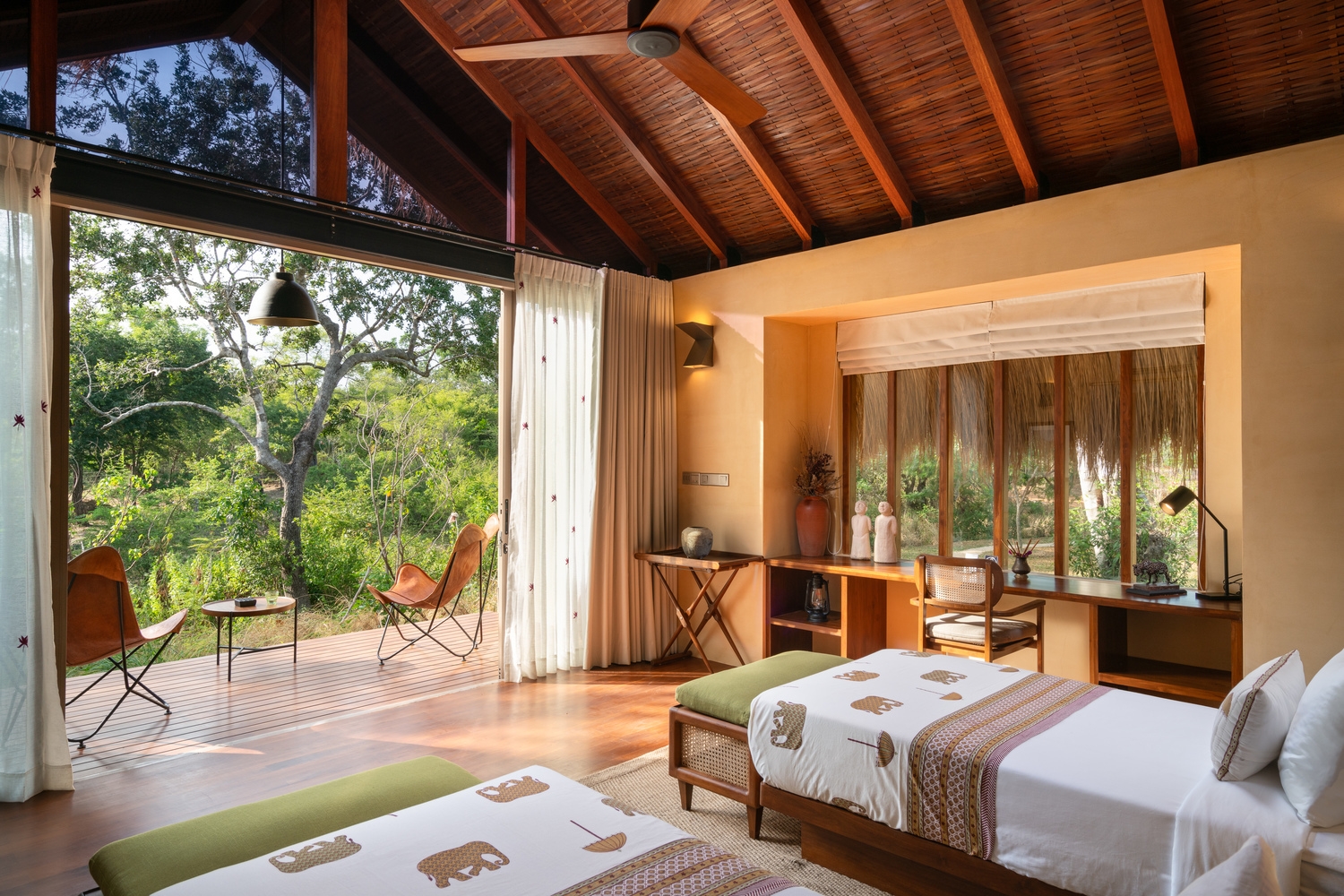 A bright bedroom with twin beds, high timber ceilings, and a large open door leading to a forest-view deck.