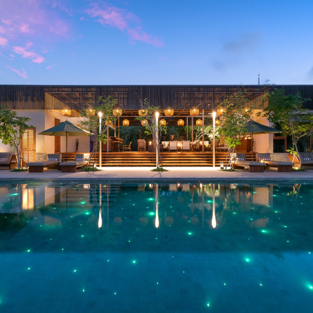 A long swimming pool at dusk with glowing green lights in the water, loungers, and a modern pavilion.