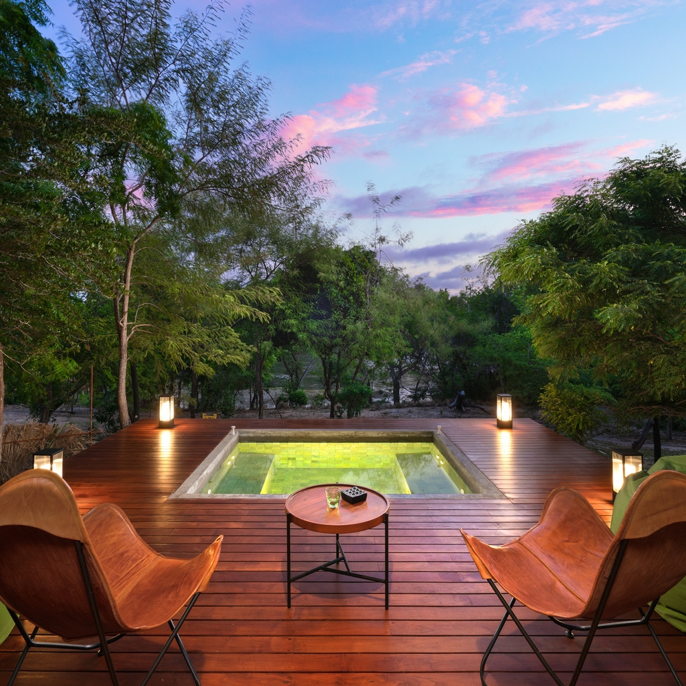 An illuminated square plunge pool set in a wooden deck with two leather chairs facing a lush forest at sunset.