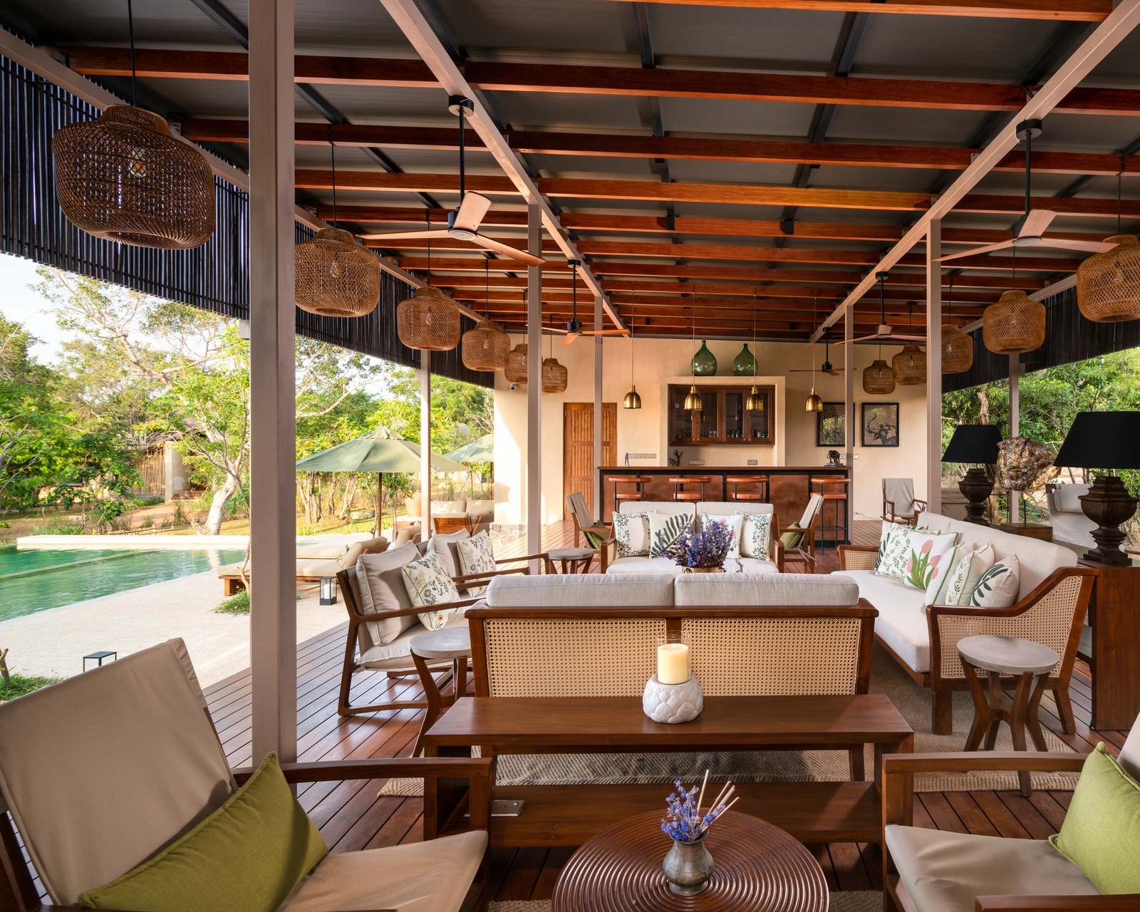 A spacious outdoor lounge with high timber ceilings, cream sofas, and large woven lanterns overlooking a garden.
