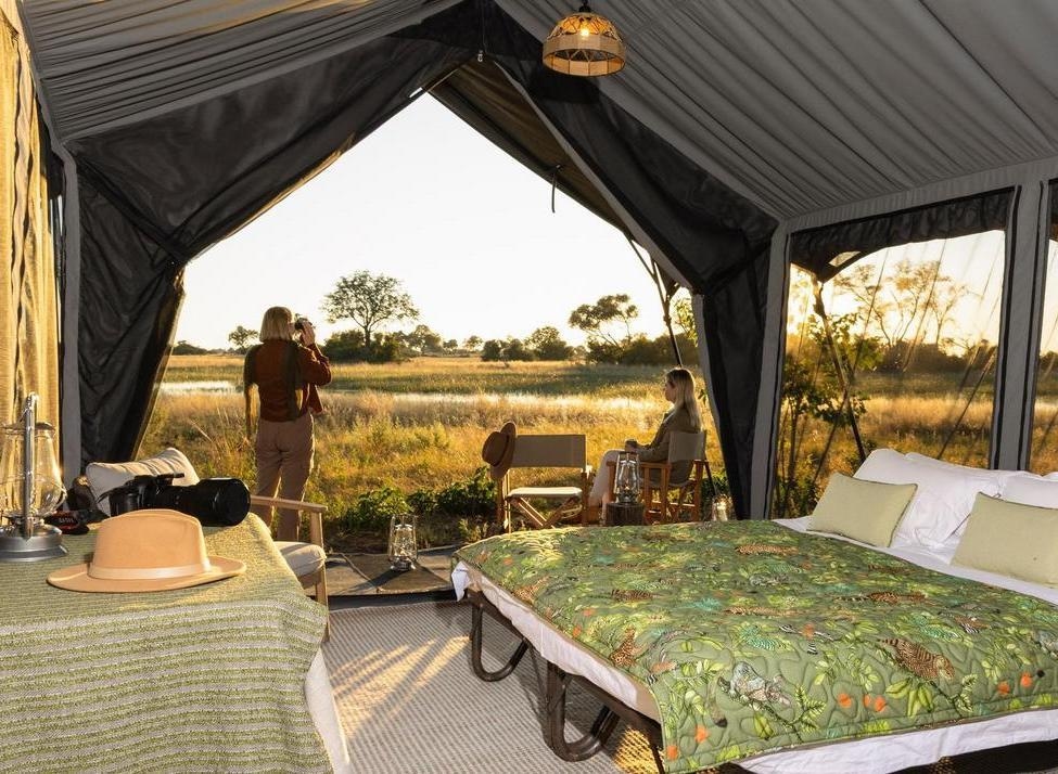 Interior of a Beagle Expeditions safari tent showing a bed and two people looking out at the savanna during sunset.