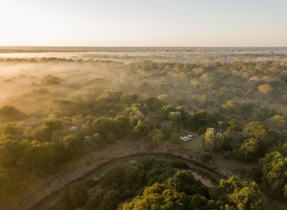 Aerial view of a safari lodge nestled in a lush forest covered in golden morning mist by a winding river.