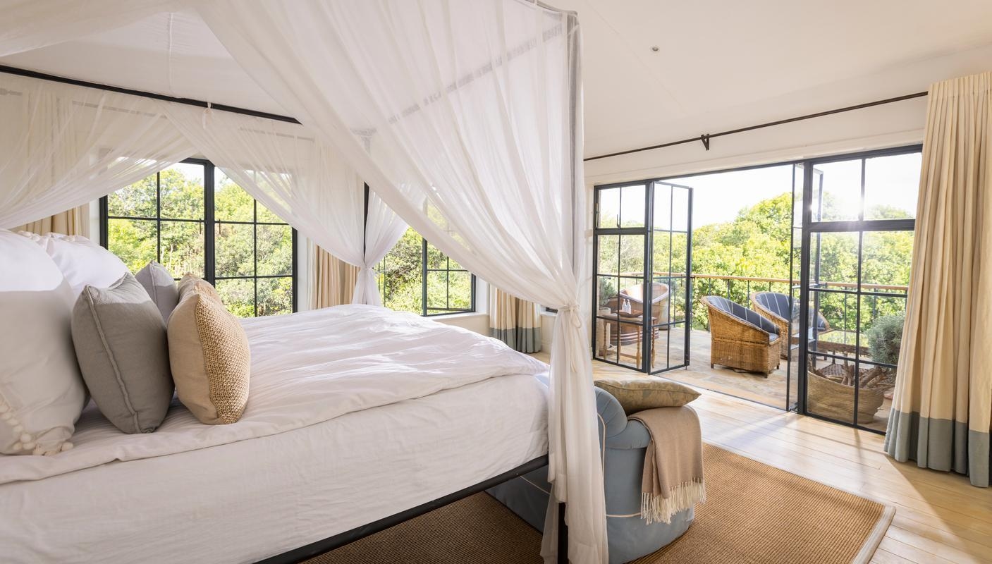 A white canopy bed in a bright room with open black-framed doors leading to a balcony and forest views.