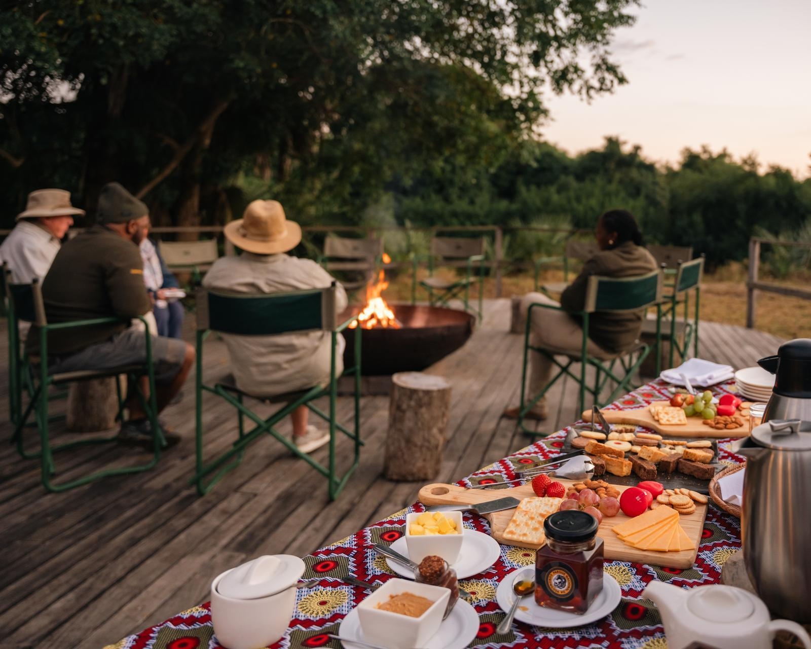 People sit by a campfire on a deck next to a table filled with fruit, cheese, and pastries at Muzimu Lodge.