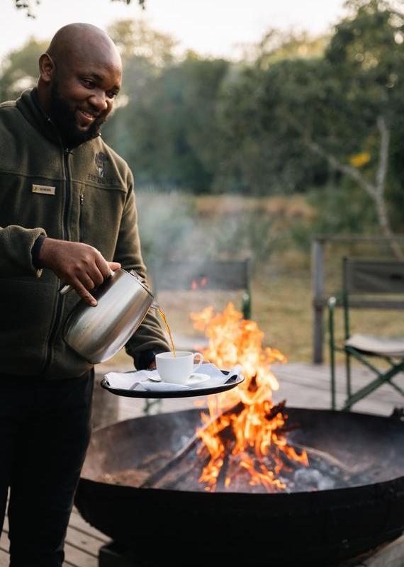 Close-up of a man pouring coffee from a silver pot into a cup beside a roaring campfire at a safari lodge.