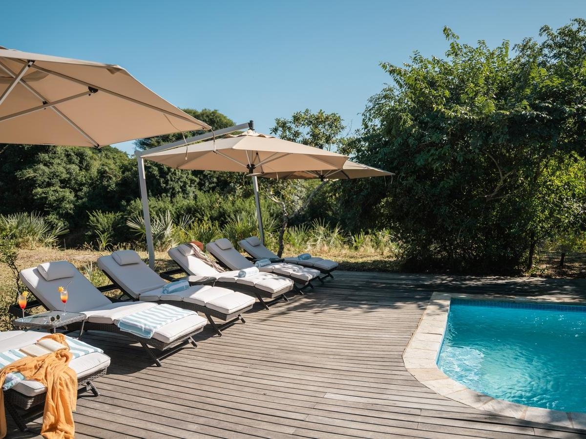 Sun loungers with towels and drinks sit on a wooden deck next to a bright blue swimming pool in a forest.