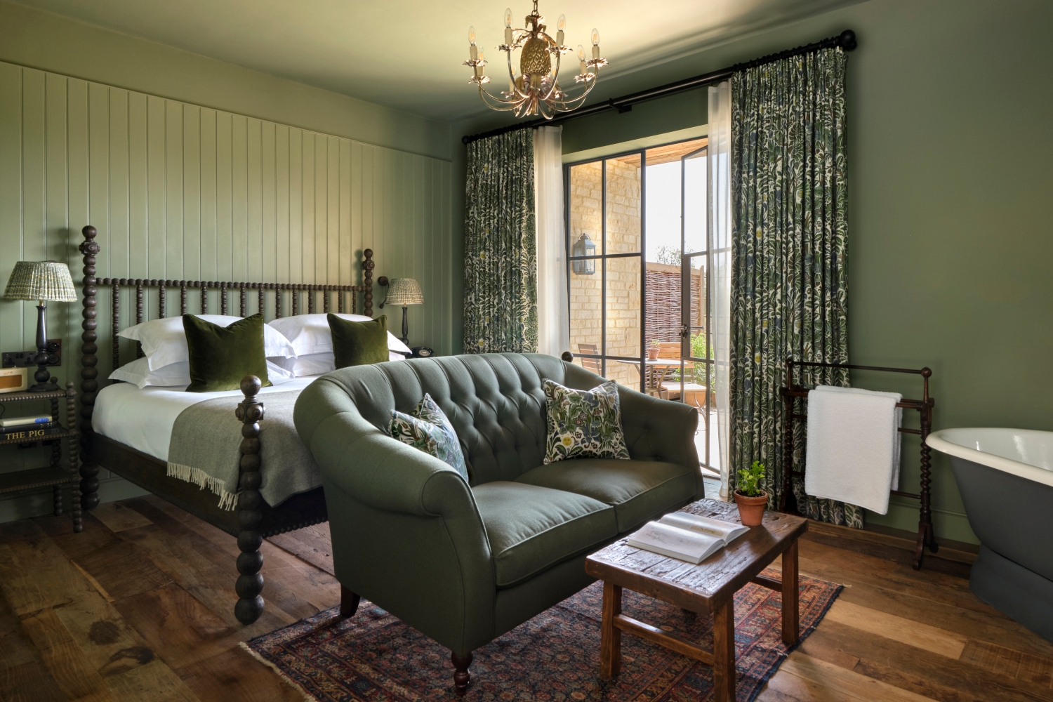 Bedroom interior with light green decor. In the foreground is a green sofa and wood coffee table in front of a bed with white sheets and green accent pillows. In the background is a large window with floor-length green curtains.