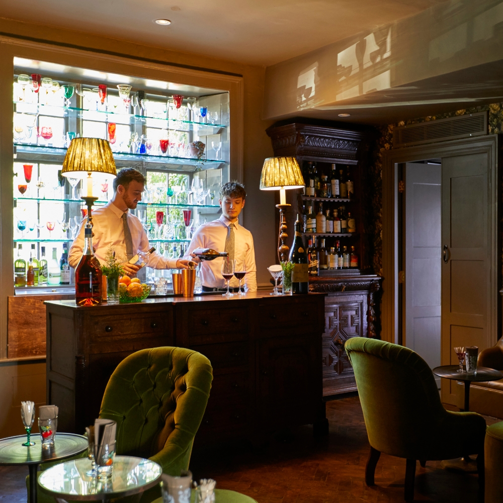 Two bartenders standing behind a small bar in front of a stained glass window. In the foreground are several plush green chairs and low bar tables, and red carpeting.