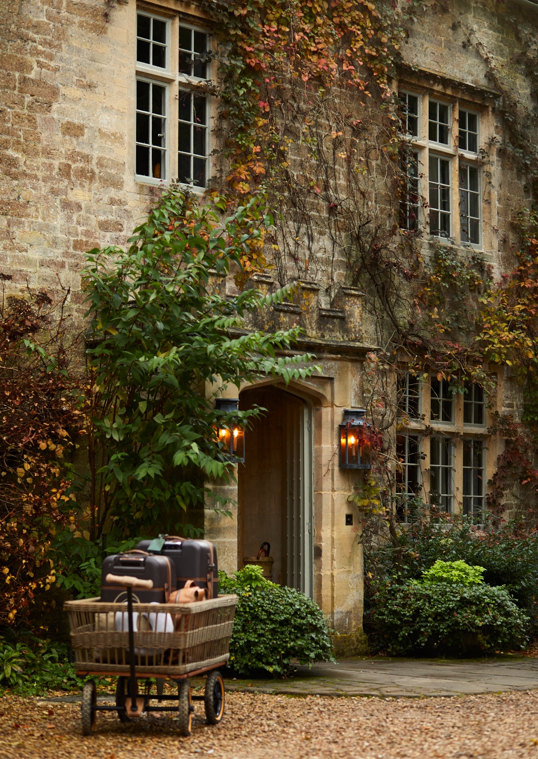 Exterior of the front entrance to the manor, with a stone facade and Georgian windows, and a door framed by two amber lamps, and a set of luggage on the gravel drive in the foreground.