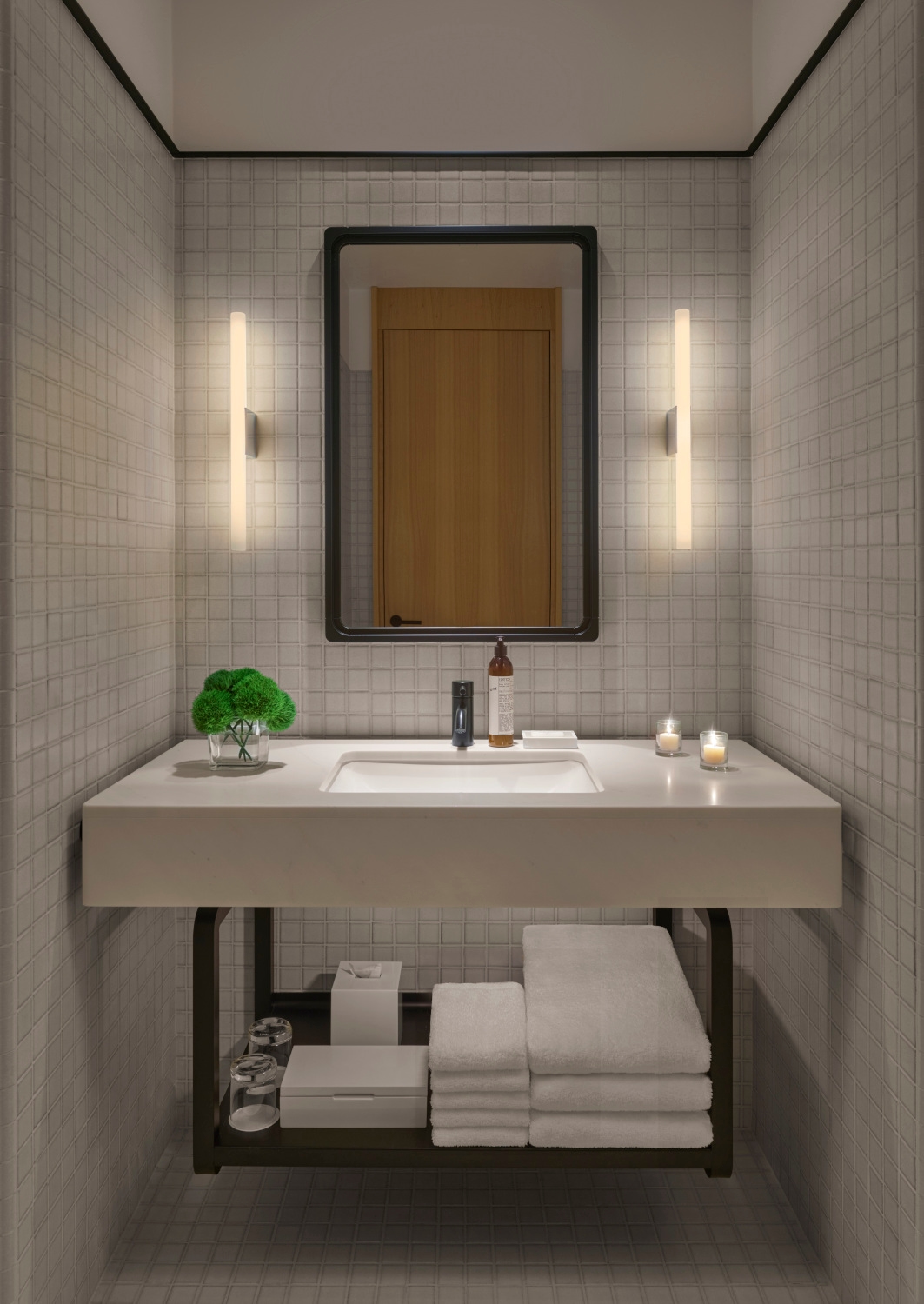 Interior of a bathroom with a vertical mirror and white tiles, and a white sink with soap bottles, lit candles and a little green decorative plant.