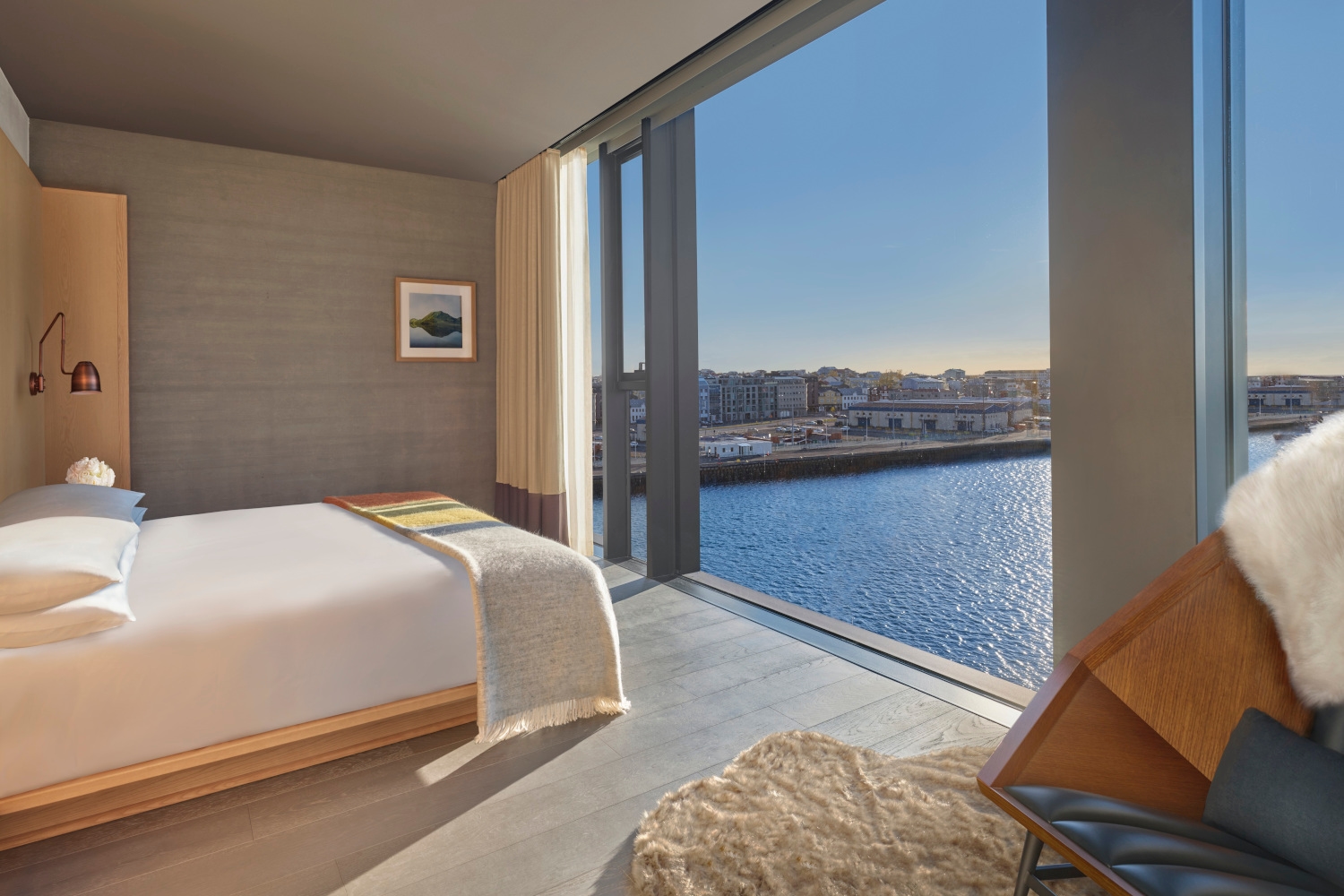Bedroom with grey walls and a bed with white and tan sheets facing a floor-to-ceiling window overlooking the harbour.