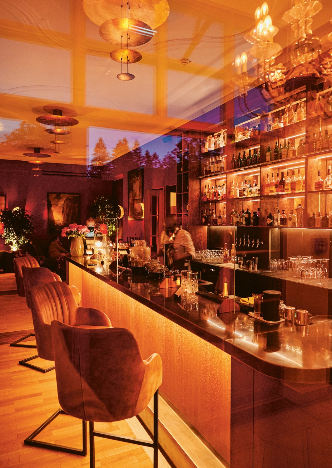 Interior of a red and brown-lit bar counter with bottles behind the counter and a row of plush bar seats along the front.