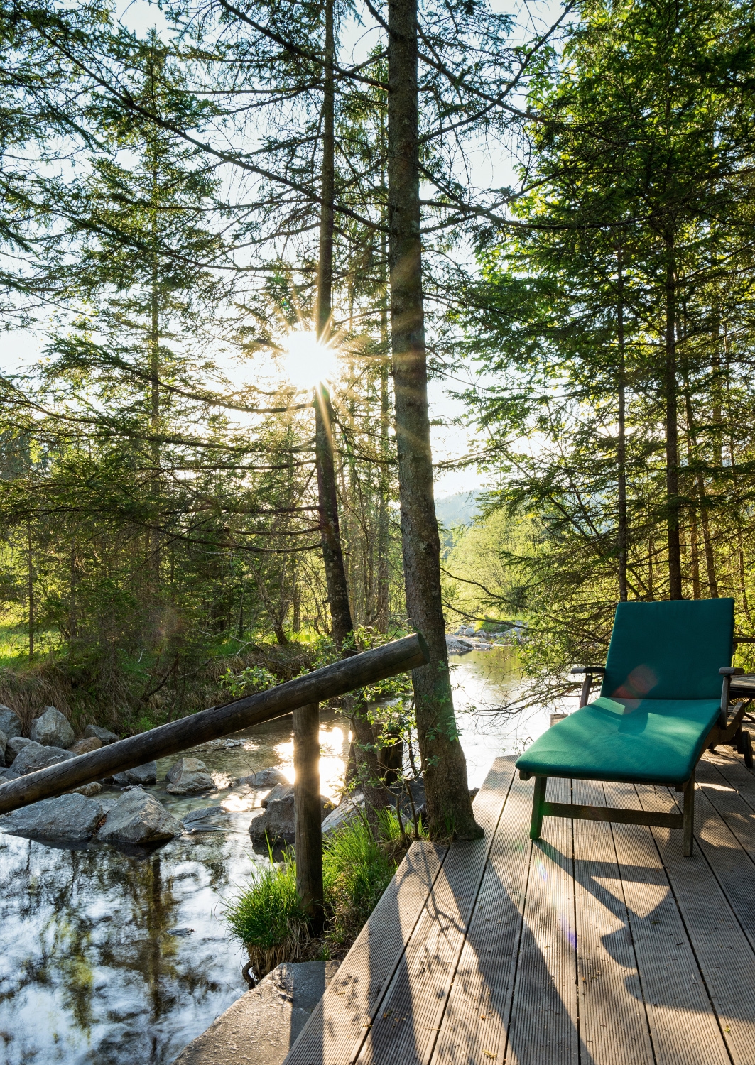 Exterior of a wooden terrace with a green lounge chair alongside a small river with sun shining through tall trees.