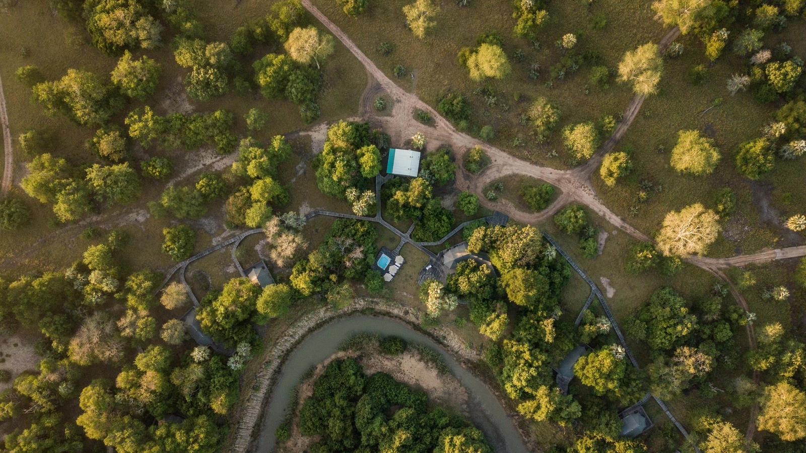 High-angle aerial shot of safari tents, a swimming pool, and wooden paths through a green landscape.