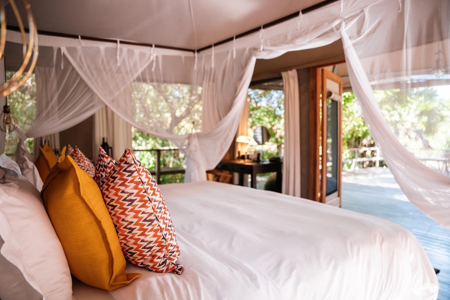 View from a bed with orange patterned pillows looking out through open tent doors toward a sunny forest deck.