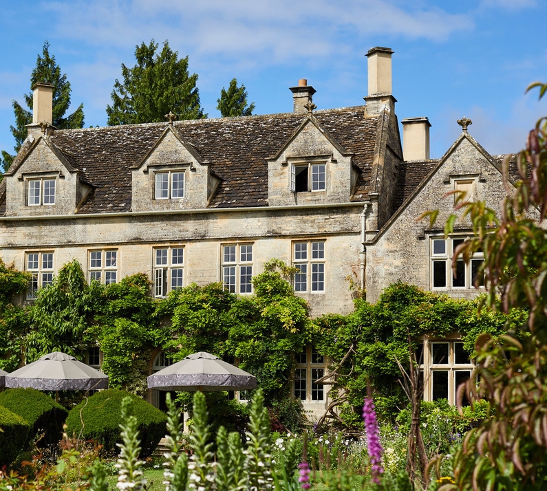 Stone manor house at THE PIG-in the Cotswolds with grey patio umbrellas and manicured gardens under a blue sky.