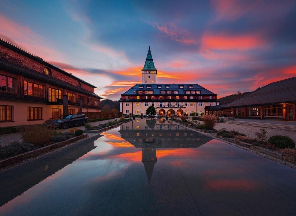 Symmetrical view of Schloss Elmau at sunset with red clouds reflected in a long courtyard pool.