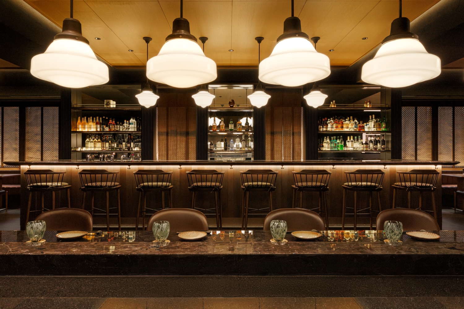 Dark wood bar and high barstools in the background with a long table in the foreground and round tiered hanging lights above.