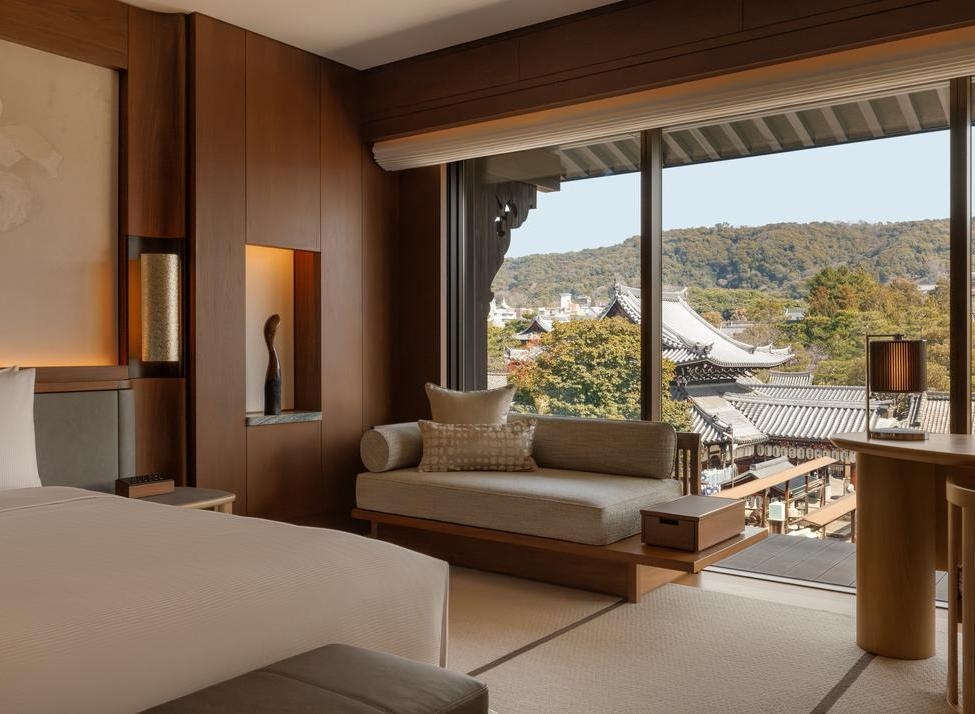 A Capella Kyoto guest room with wooden walls, a daybed, and a large window looking out onto a traditional temple complex.