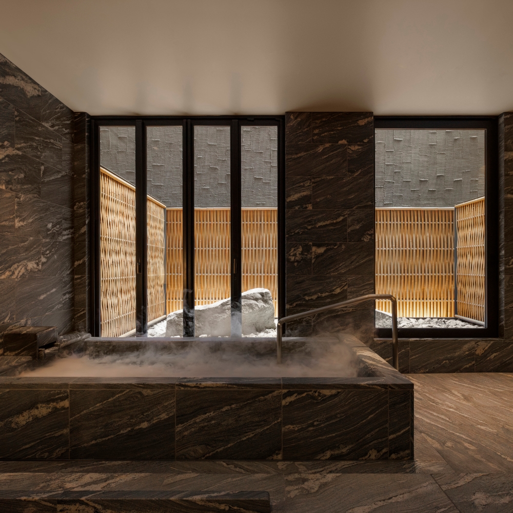 Private onsen tub steaming with water; the bathroom has dark tiles and there are windows overlooking an outdoor bathing terrace surrounded by a bamboo or rattan fence.