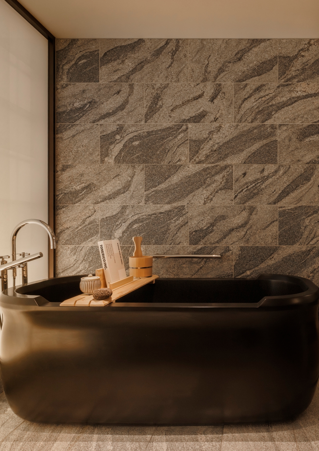 Dark stone soaking bathtub with a tray of toiletries in a room with grey stone tiled walls.