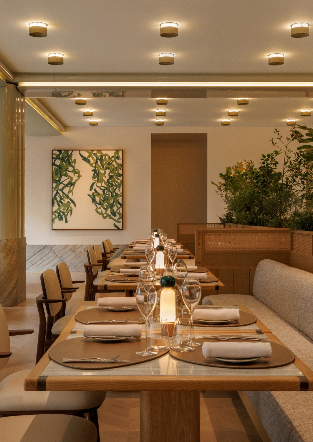 View down a long dining table set with plates and wine glasses. In the background are walls and banquettes and artwork with muted tones.