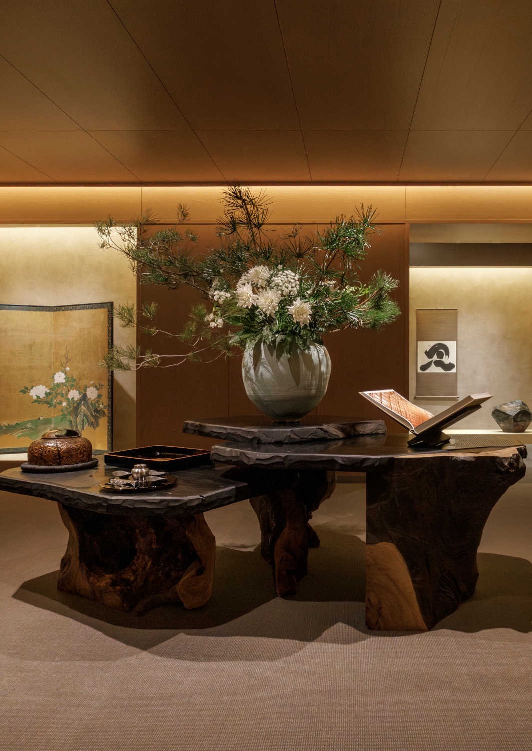 Lobby reception with a welcome table made of wood fossils on top of which are a large vase with a floral arrangement and a decorative book open on a stand.