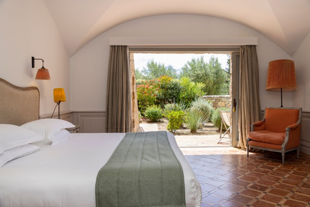 A hotel room with a bed, armchair and sliding doors open to a sunny garden