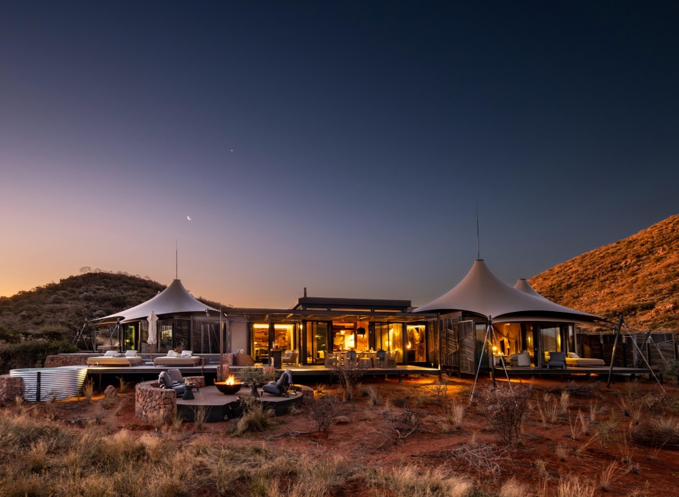 Exterior view of a tented camp at dusk, with three pointed tents surrounding a glass and open-air lobby with a large deck and in the foreground a table, chairs and firepit, all set in bushland with red soil and short grass.