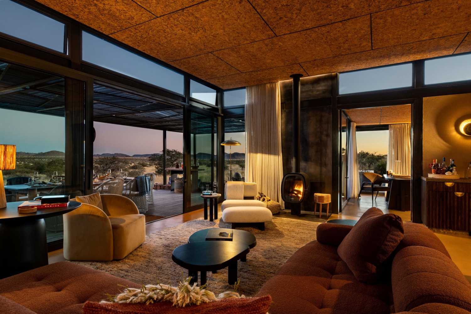 Interior of a villa living room with dark sunset outside, and inside a selection of upholstered chairs, loungers and sofas, a black coffee table and rugs, with huge windows looking out to a deck and the bush beyond.