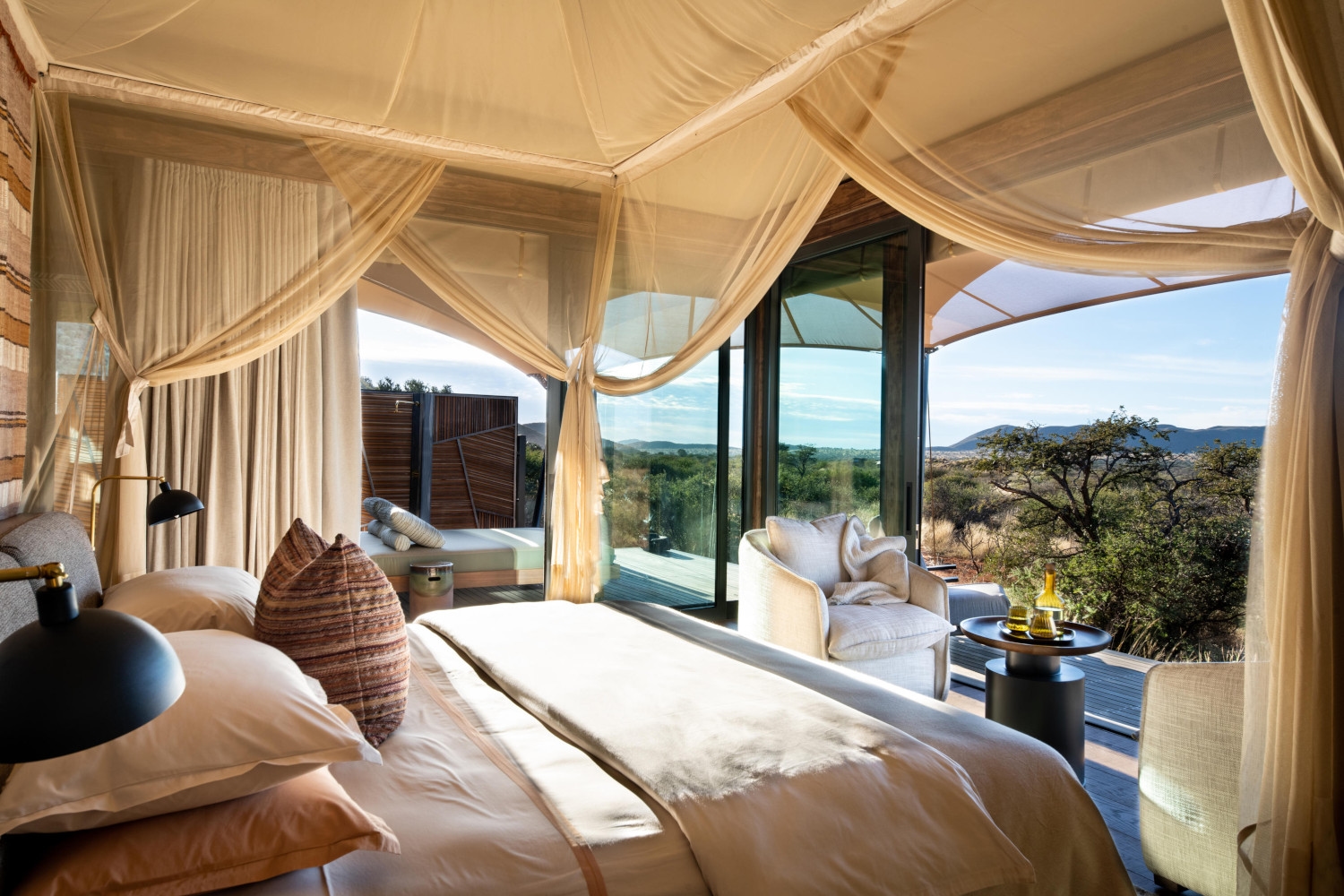 Double bed surrounded by gathered mosquito nets and dressed in beige and brown bedding. The room has picture windows on two sides looking onto the bush, and beside the bed a white lounge chair and small table.