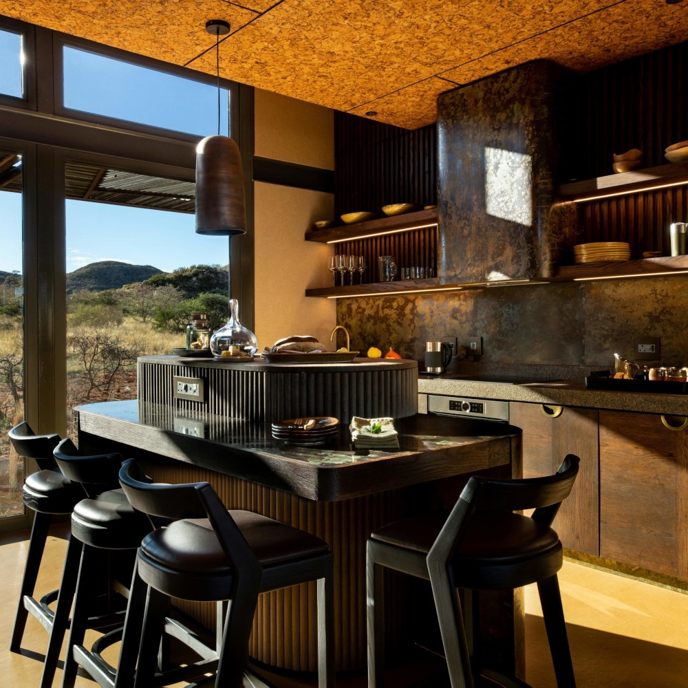 Kitchen with dark stone backsplash and cabinets, and a centre console with four black stools, and a large window looking out to the bush.