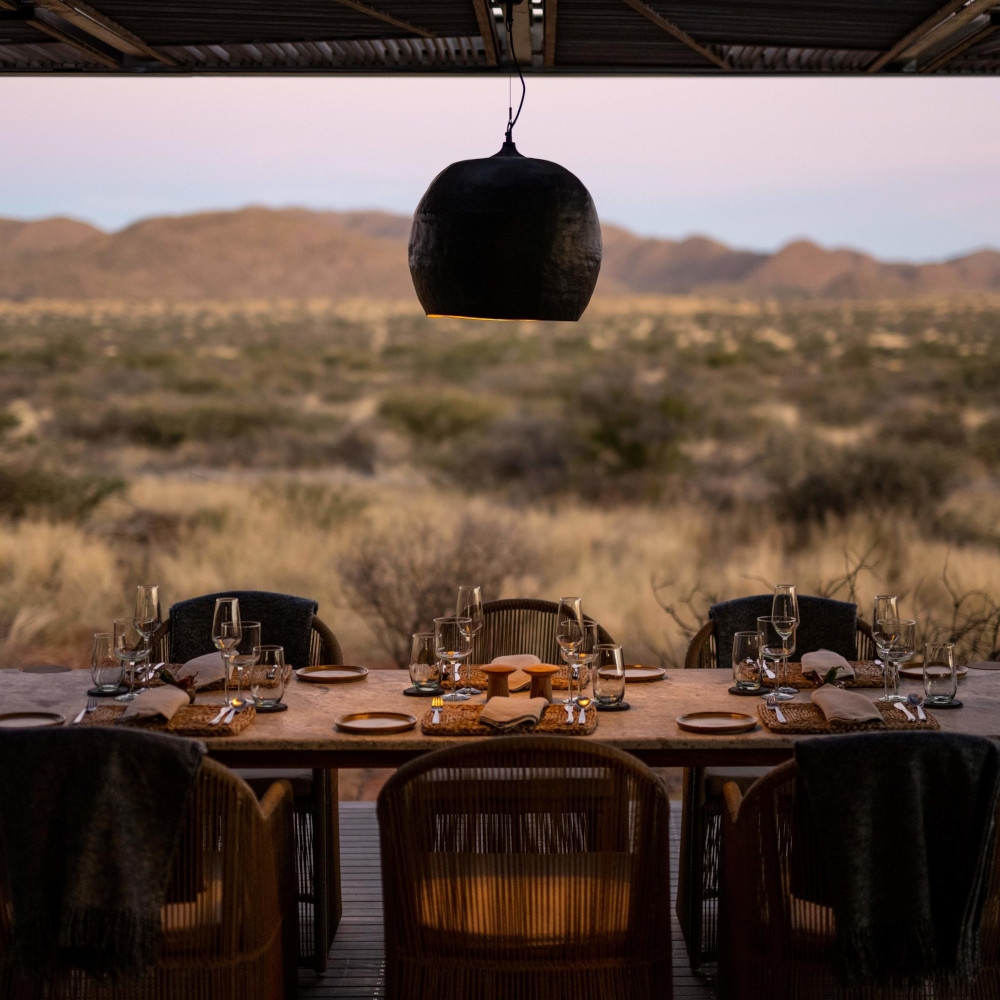 Six chairs at a table set with dinner plates and wine glasses with a round black hanging lamp shade above and beyond an open-air view of the bush and mountains.