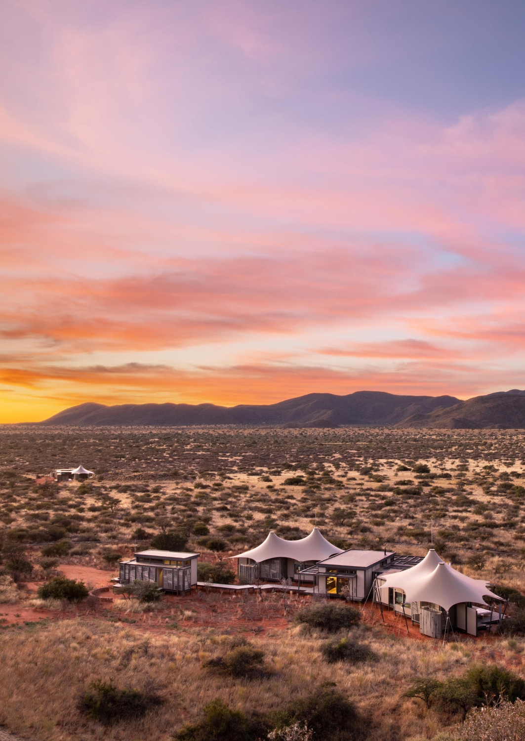 Aerial shot of two tented villas, one in the foreground and one in the distance, set in a wide savannah with low bushes and mountains in the background, all covered by a pink sunset.