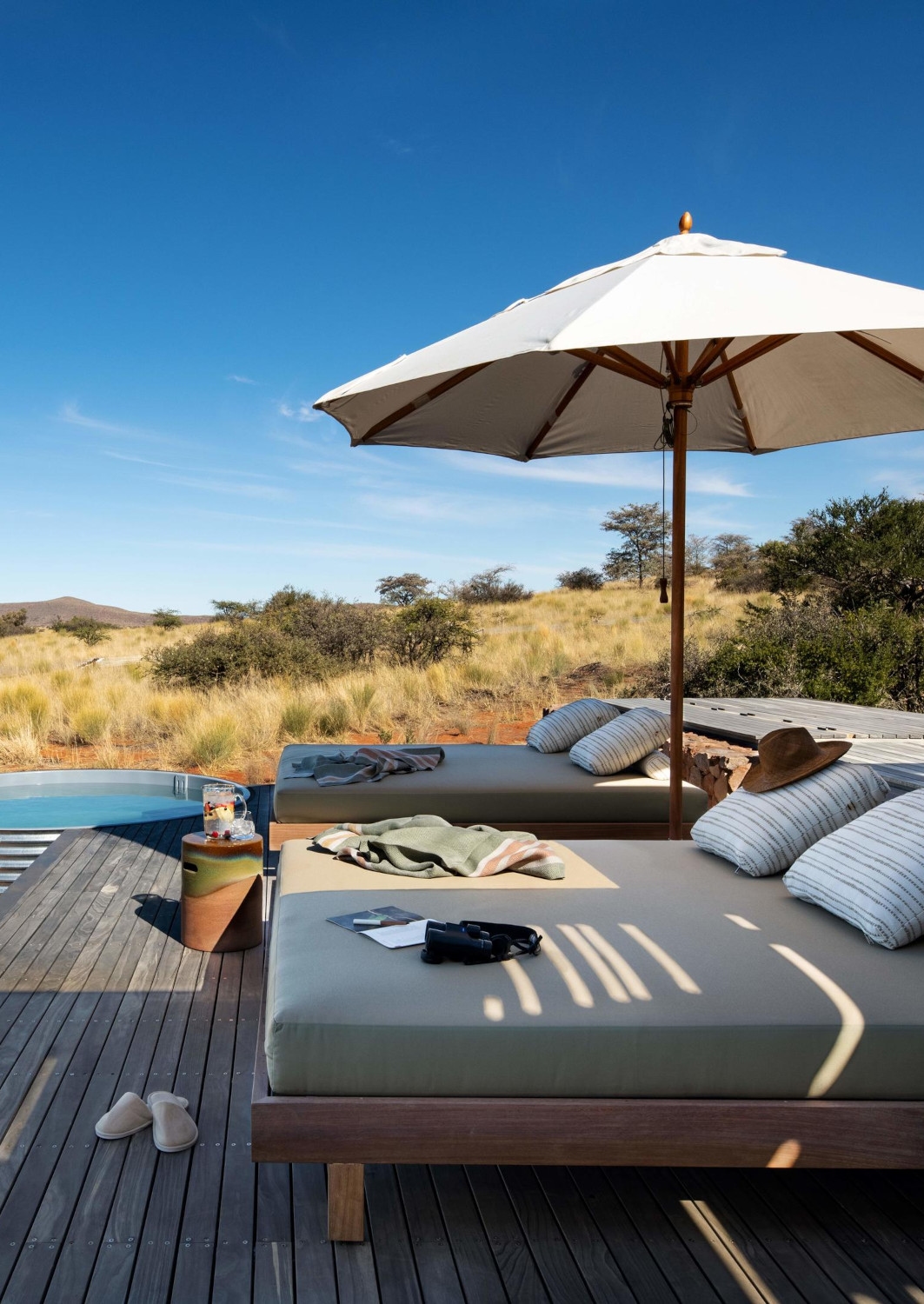 Two sun loungers beneath a white umbrella sitting on a wooden deck next to a pool. The loungers have assorted accessories on them, including a sun hat, books, slippers, towels, drinks. The bush is just beyond the pool.